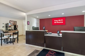 Un espace d’accueil d’hôtel avec un mur rouge, un panneau indiquant « Wi-Fi gratuit dans toutes les chambres », une machine à café et un coin salon avec des chaises et une table.