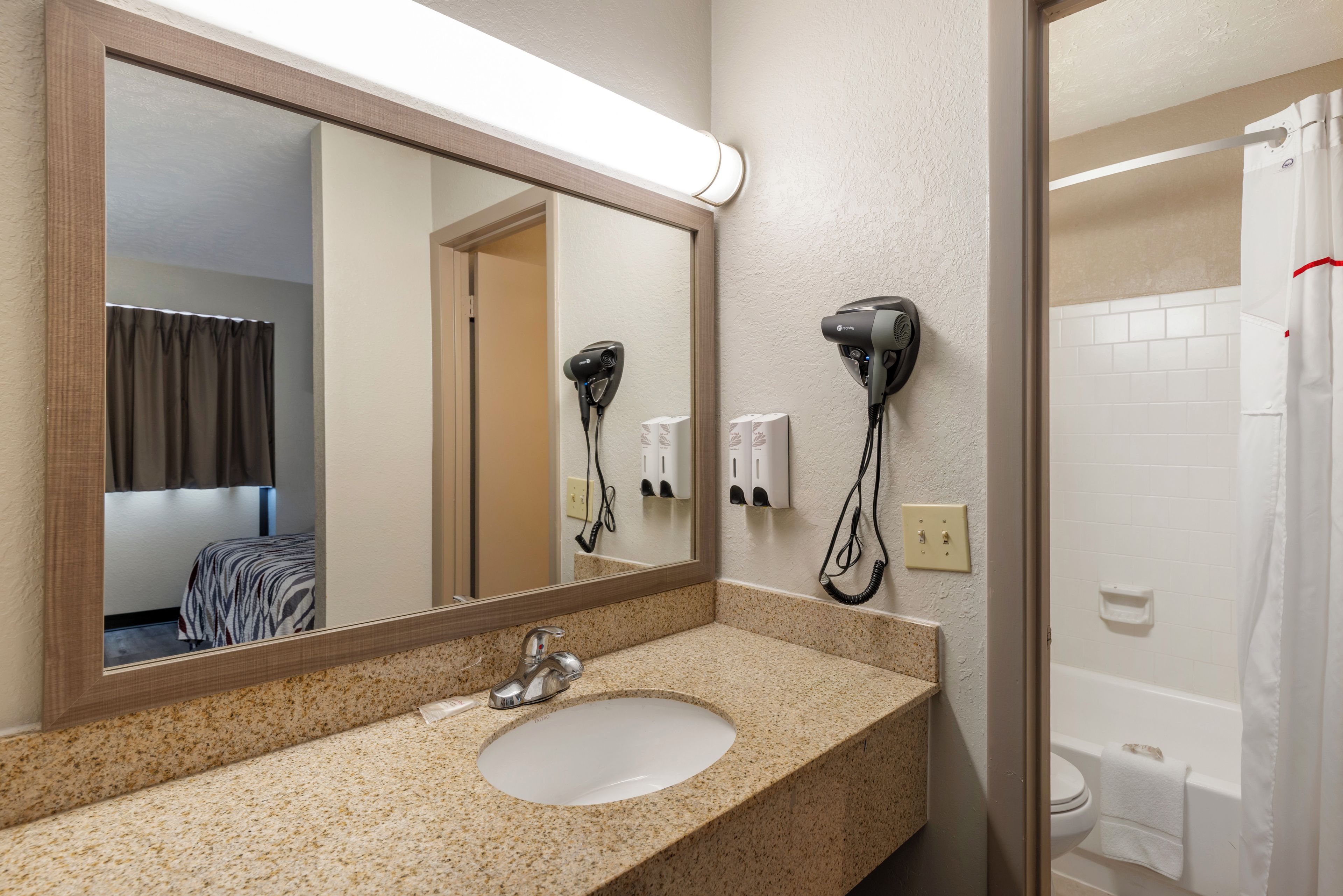A hotel bathroom with a sink, mirror, hair dryer, and a shower area.