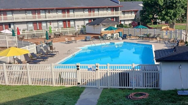 Une piscine entourée de chaises longues et de parasols, le tout délimité par une clôture blanche, devant un motel comprenant de nombreuses chambres.