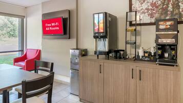 Une petite cuisine moderne équipée d’une machine à café, d’un réfrigérateur, d’un téléviseur affichant le message « Wi-Fi gratuit dans toutes les chambres » et d’une chaise rouge.