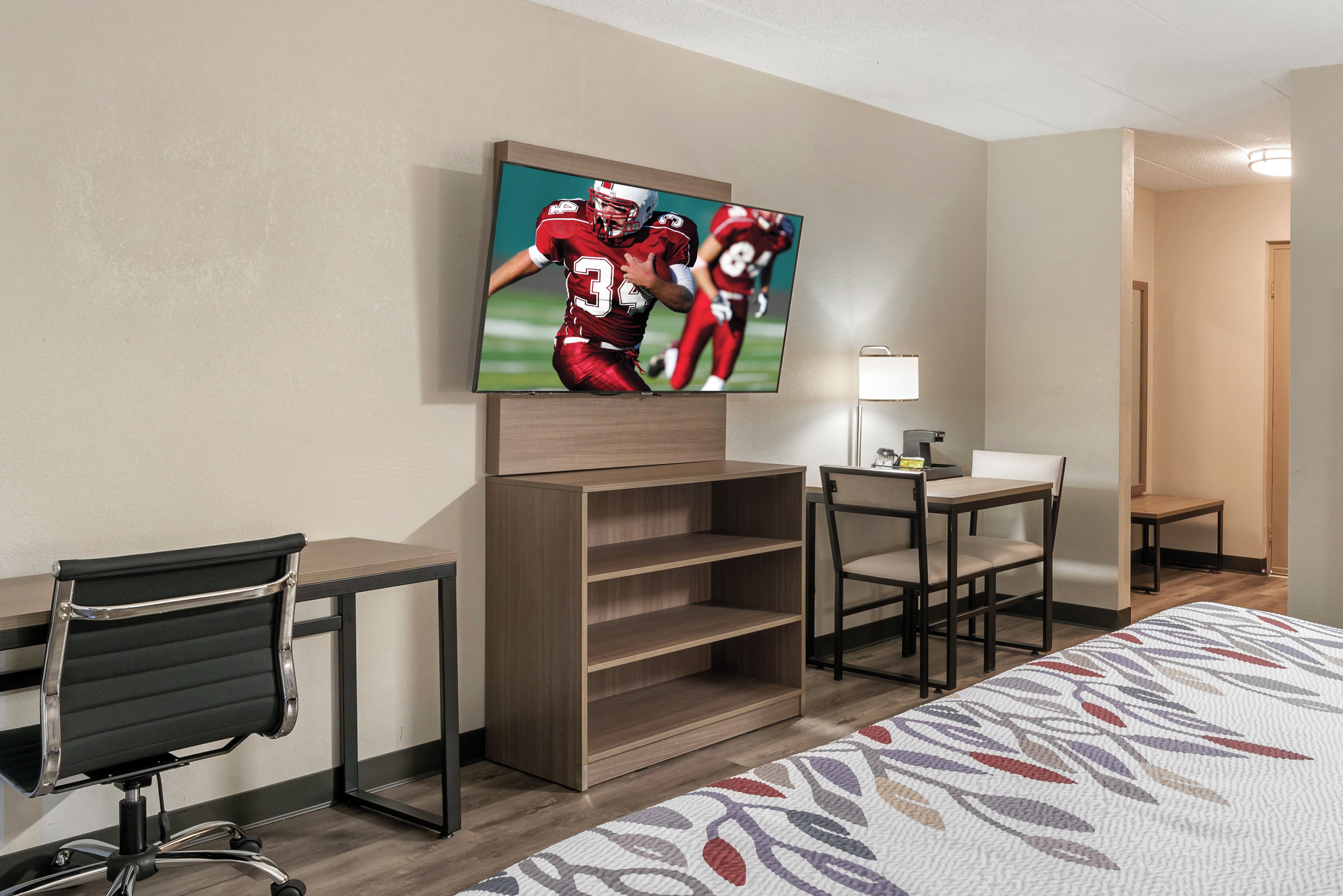 A hotel room with a bed, a desk, a chair, a TV mounted on the wall, and a small table with a lamp.