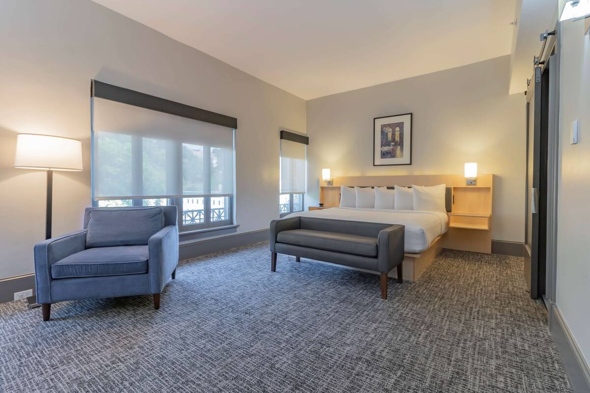 A hotel room with a bed, two armchairs, a standing lamp, and a large window.