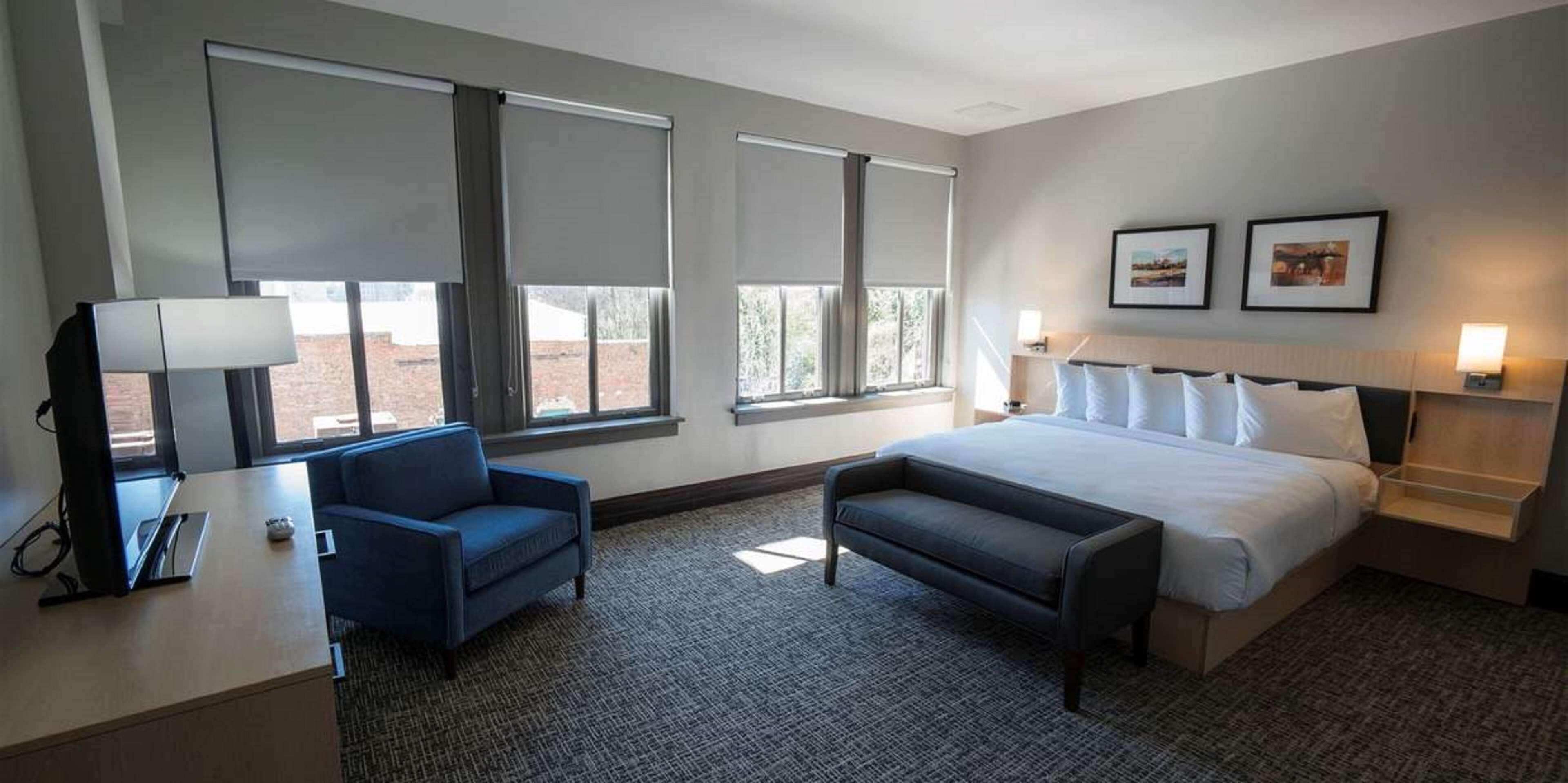 Une chambre d’hôtel avec un lit, un bureau avec une lampe, une chaise et deux fenêtres équipées de stores.