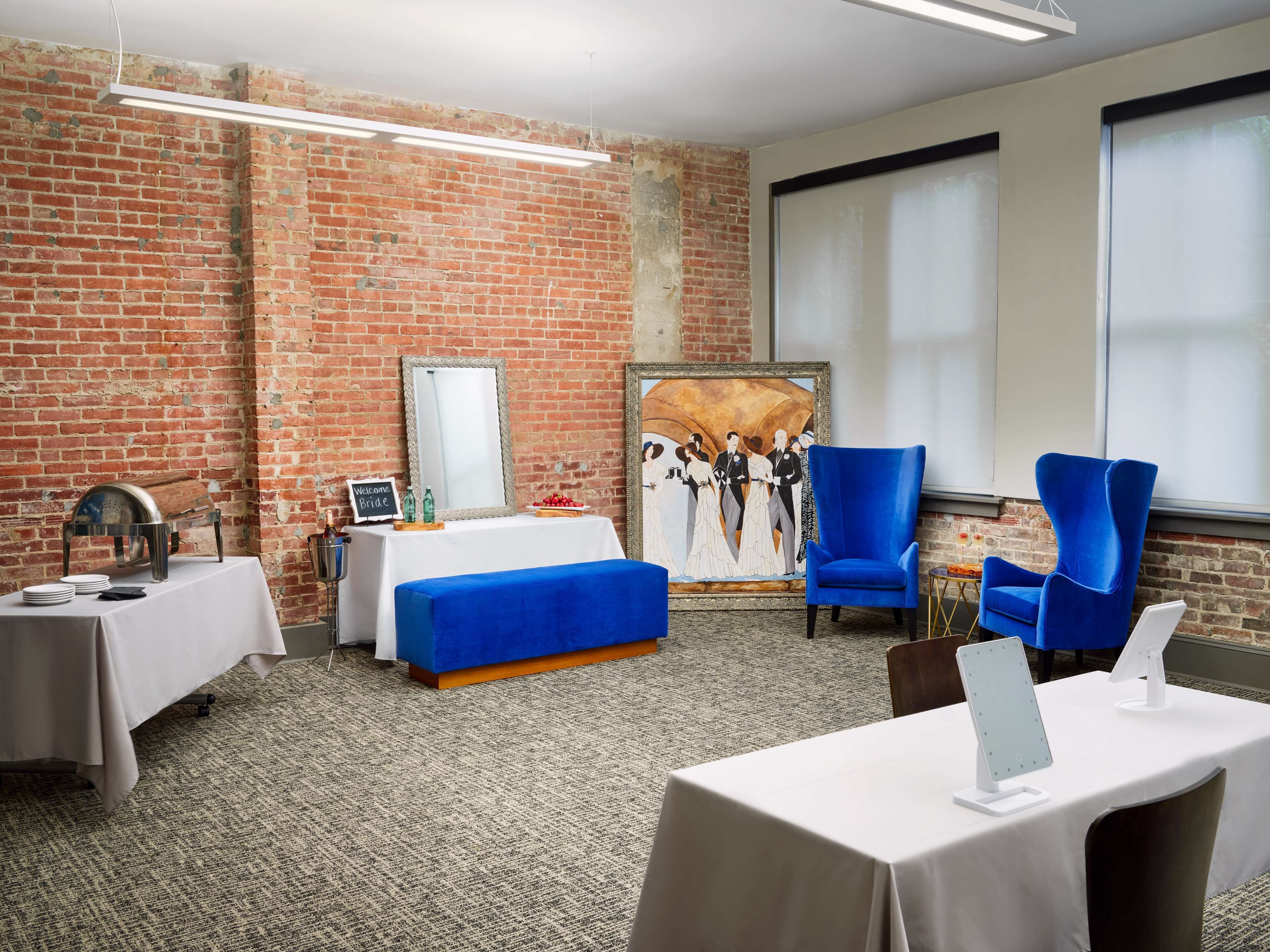 Ein Hotelzimmer mit Ziegelwand, einem großen Gemälde, blauen Stühlen, einem Bett und einem Tisch mit einem Laptop.