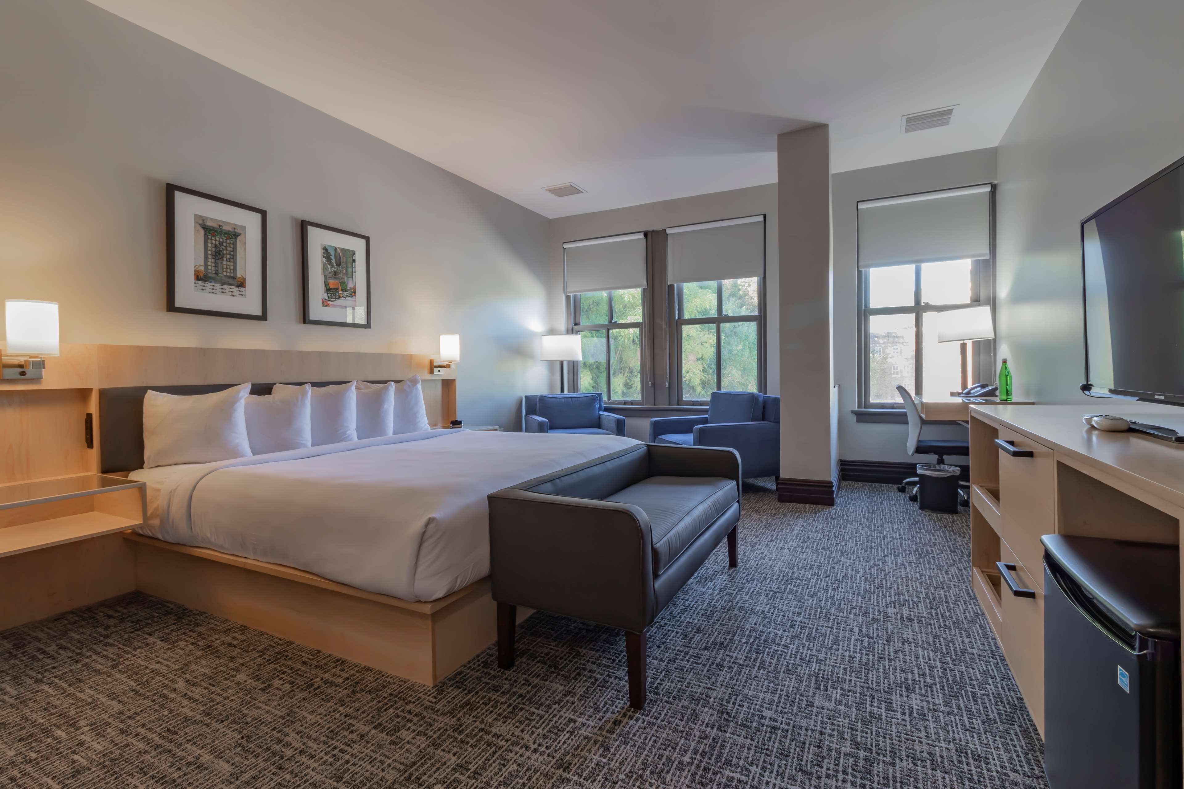 A hotel room with a large bed, a desk, two chairs, a television, and framed artwork on the wall.