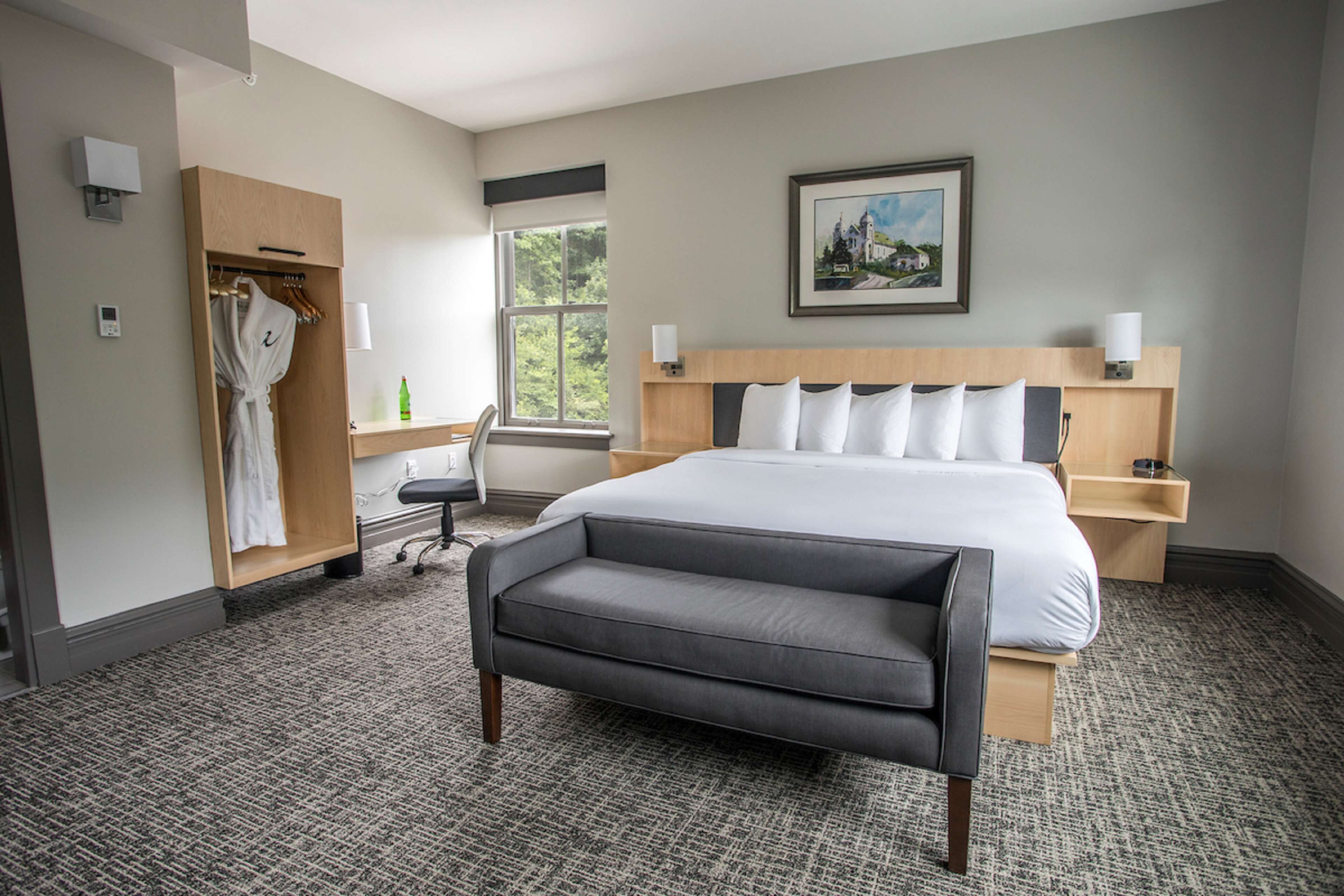 A hotel room with a large bed, a bedside table with a lamp, a wardrobe, a desk with a chair, and a window with a view of trees.