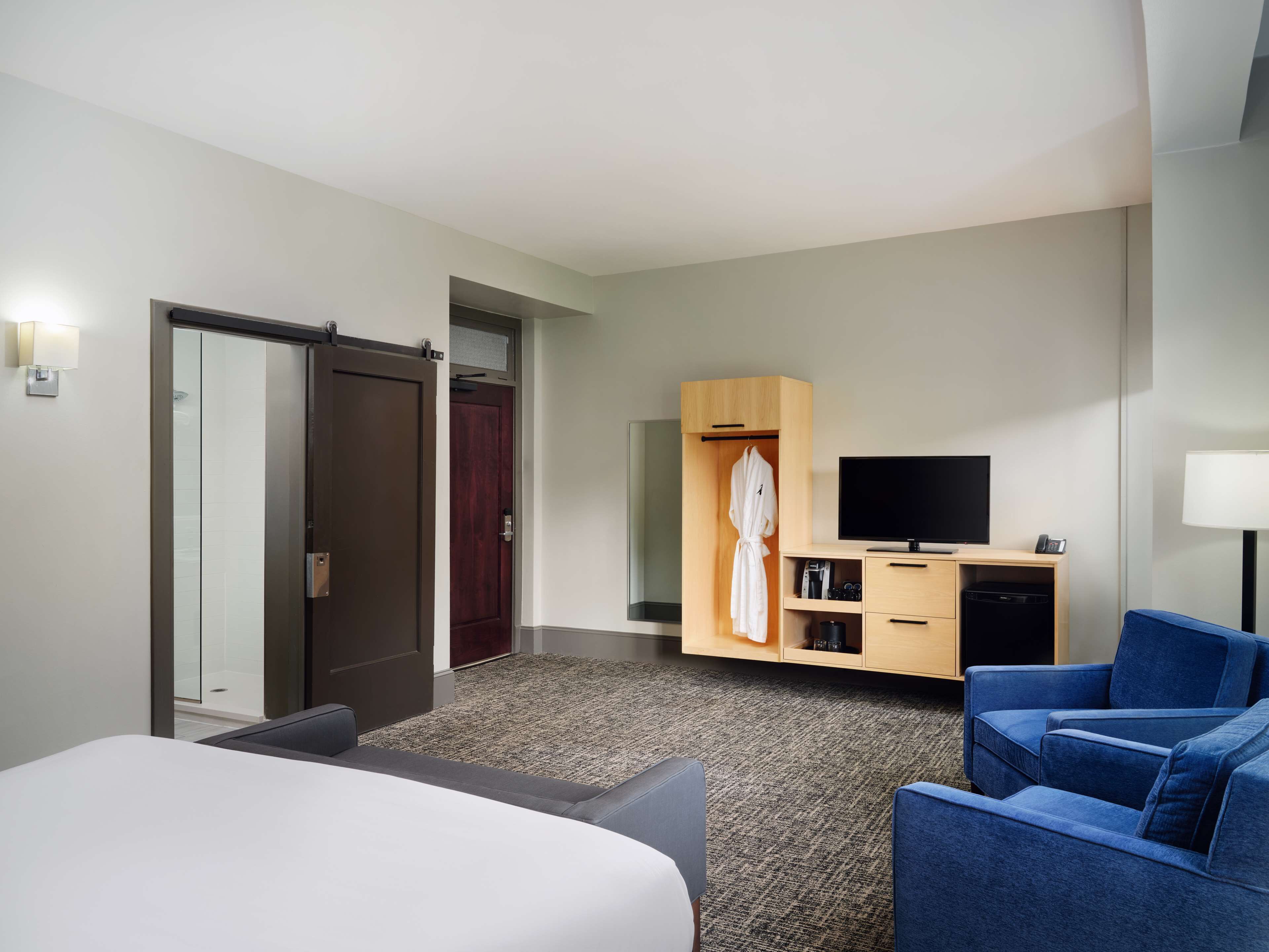 A hotel room with a bed, a seating area with a blue sofa, a TV, a wooden wardrobe, and a robe hanging inside.