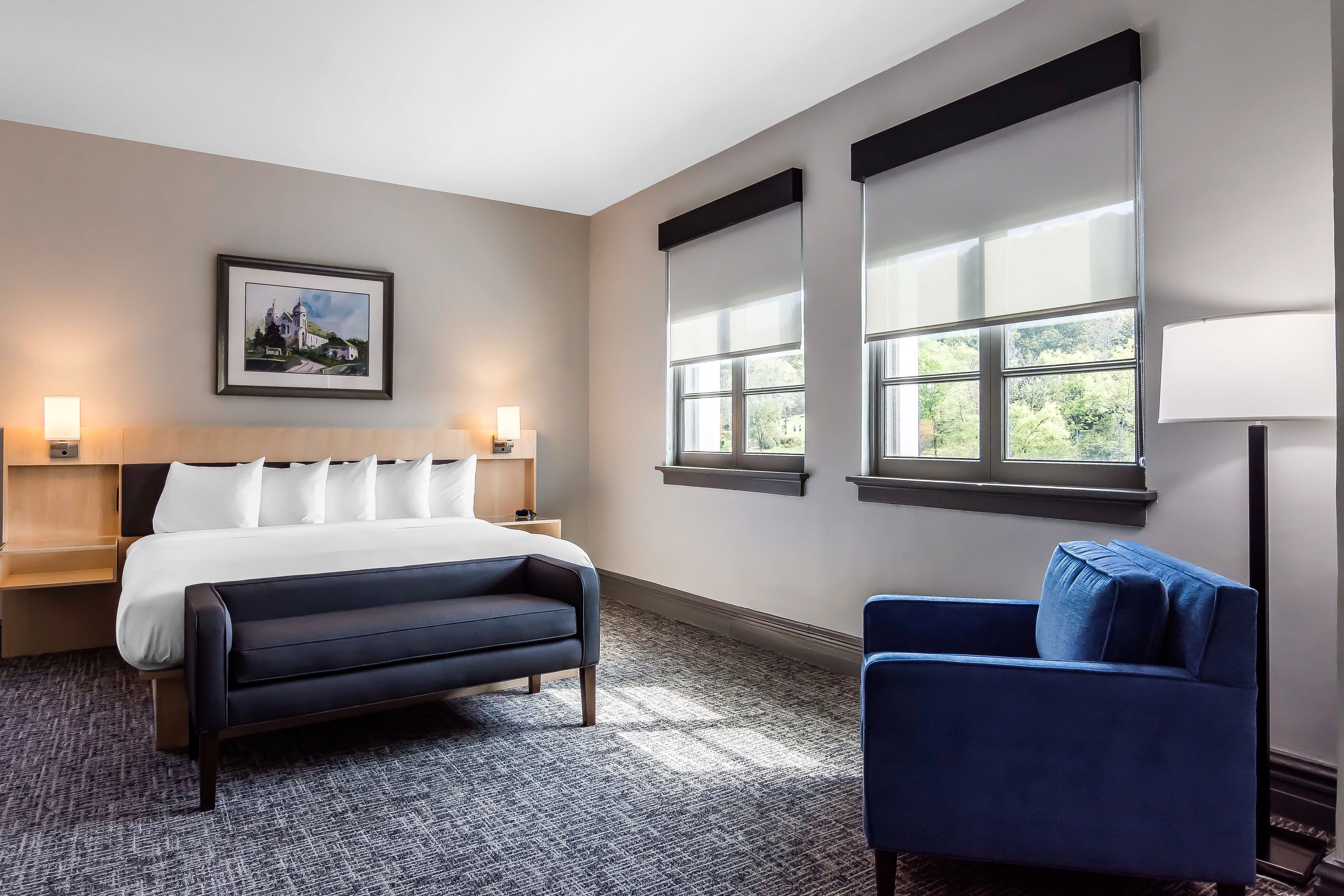 Une chambre d’hôtel avec un lit, un fauteuil bleu, deux tables de chevet avec des lampes, un tableau encadré au mur et de grandes fenêtres.