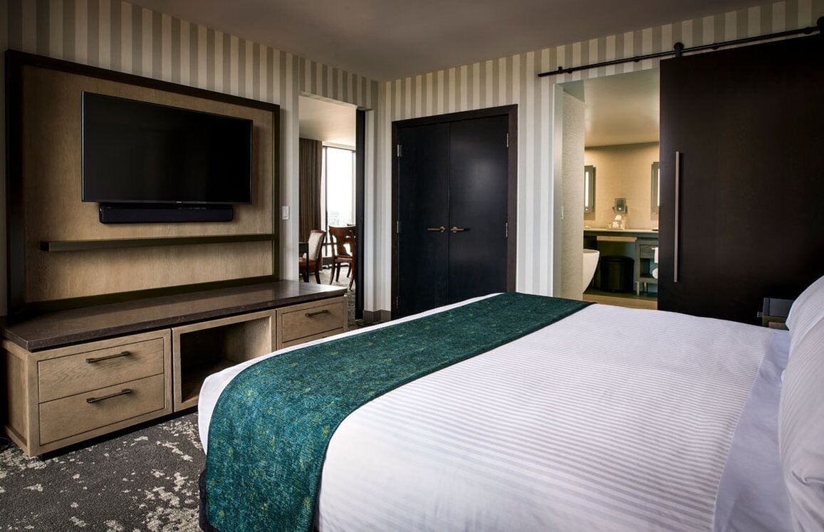 Habitación de hotel con cama, televisor montado en la pared, escritorio con cajones, una puerta y baño visible a través de una puerta abierta.