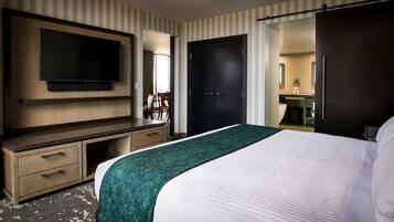 Habitación de hotel con cama, televisor montado en la pared, escritorio con cajones, una puerta y baño visible a través de una puerta abierta.