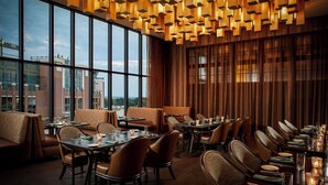 A modern dining area with high ceilings, wooden geometric light fixtures, and large windows offering views of the cityscape.