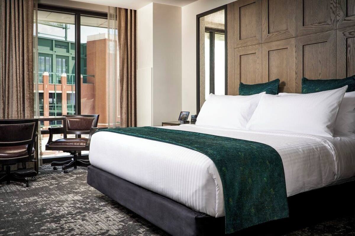 Habitación de hotel con una cama grande, un escritorio y una silla. Hay una ventana con cortinas y vistas a edificios.