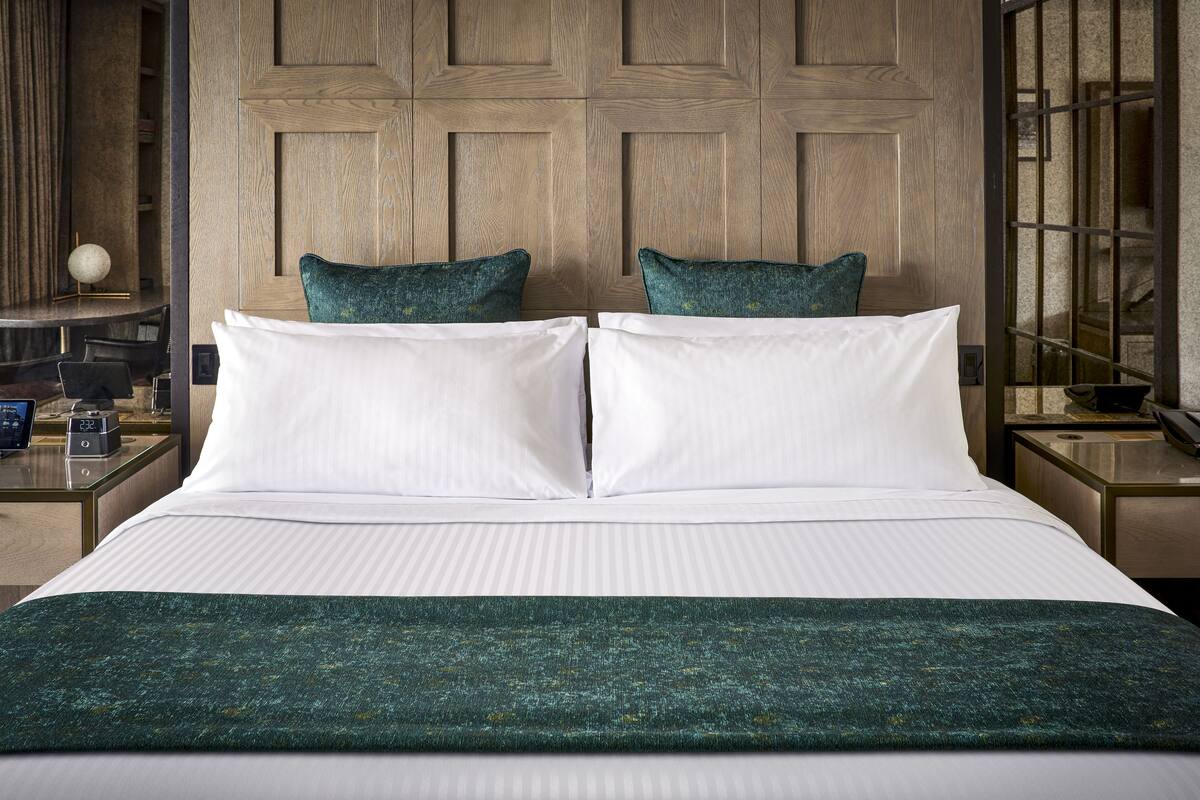 Cama doble con sábanas blancas y dos almohadas color turquesa, frente a un cabecero de madera con diseño geométrico.