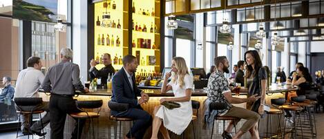 A spacious bar area with high ceilings and large windows, featuring a variety of seating options and a well-stocked bar counter.