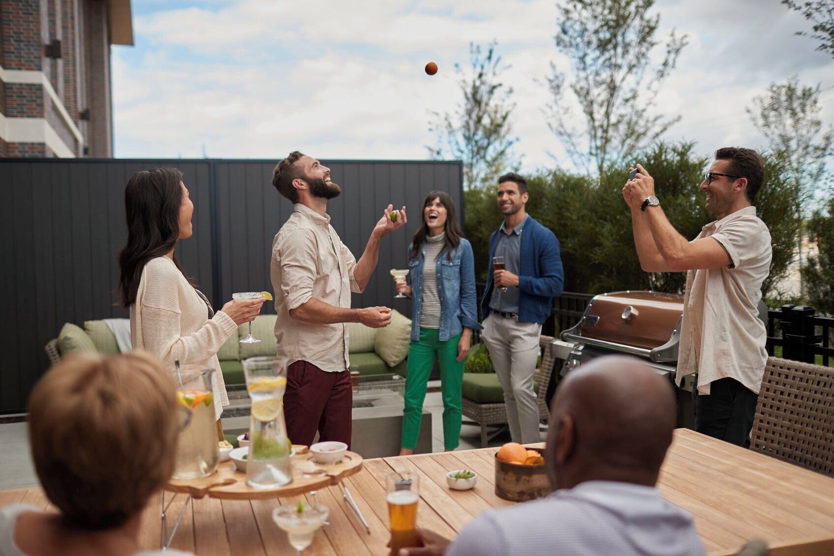 Un grupo de personas al aire libre, algunas sosteniendo bebidas, con una parrilla y una mesa de comedor con comida y bebidas al frente.