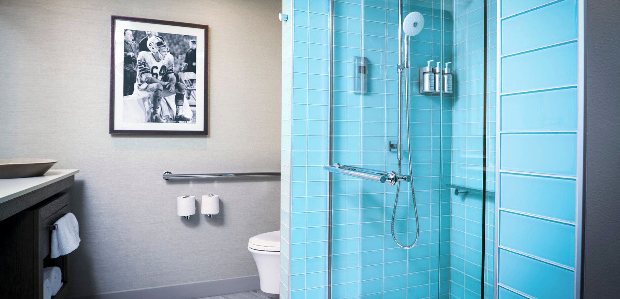 A bathroom with a blue-tiled shower, a toilet, a sink with a mirror, and a framed photograph on the wall.
