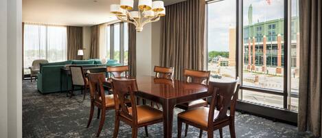 Habitación de hotel con una mesa de comedor, sillas, un sofá y un amplio ventanal con vistas a un edificio.
