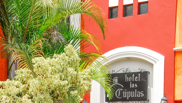 A hotel entrance with a red facade, a large potted plant, and a sign reading "hotel las Cúpulas." - Raintree`s Las Cupulas Oaxaca (San Felipe del Agua)