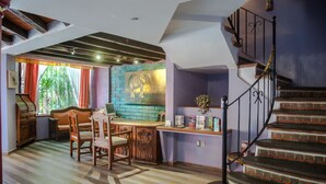 A dining area with a wooden table and chairs, a brick wall with a mural, and a staircase with decorative railings. - Raintree`s Las Cupulas Oaxaca (San Felipe del Agua)