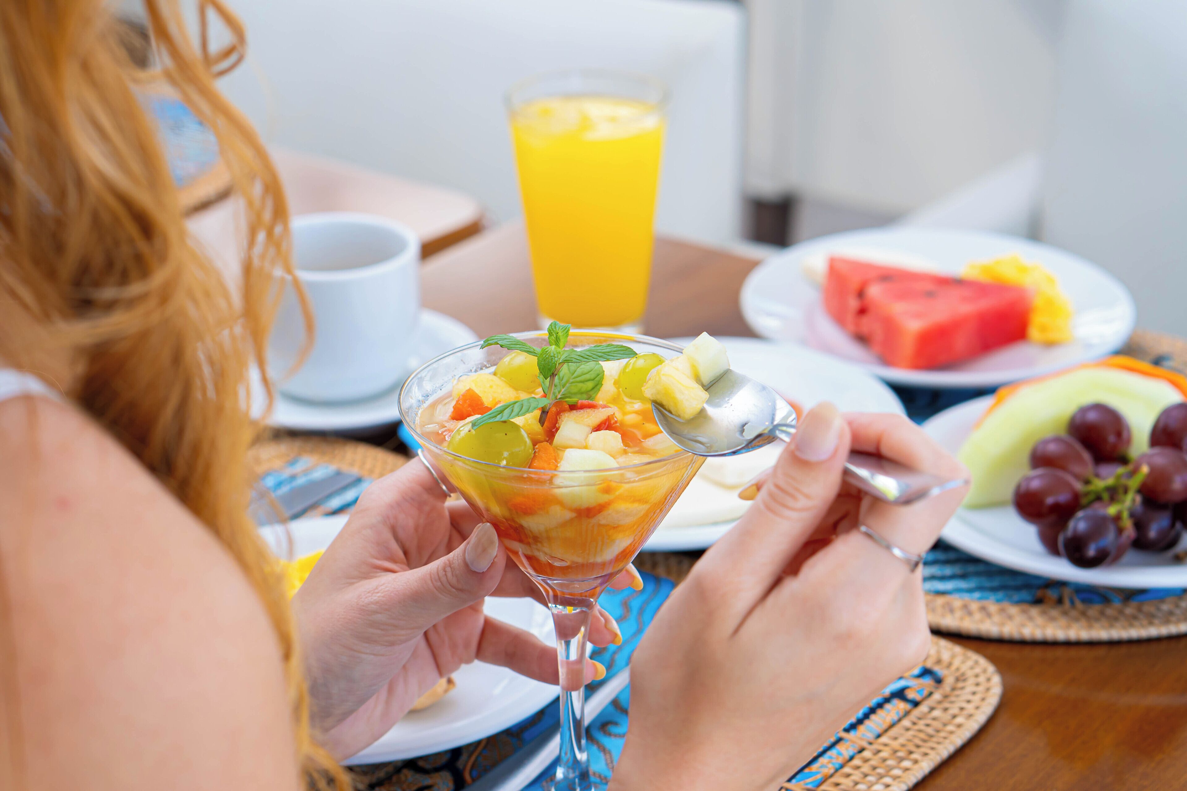 Una persona sosteniendo una ensalada de frutas en un vaso, un vaso de jugo de naranja y platos con sandía y uvas.