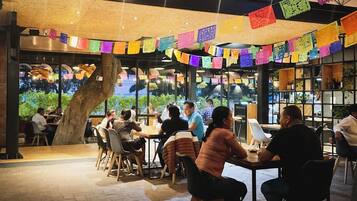 An indoor dining area with tables and chairs, a large tree trunk, and colorful paper decorations.