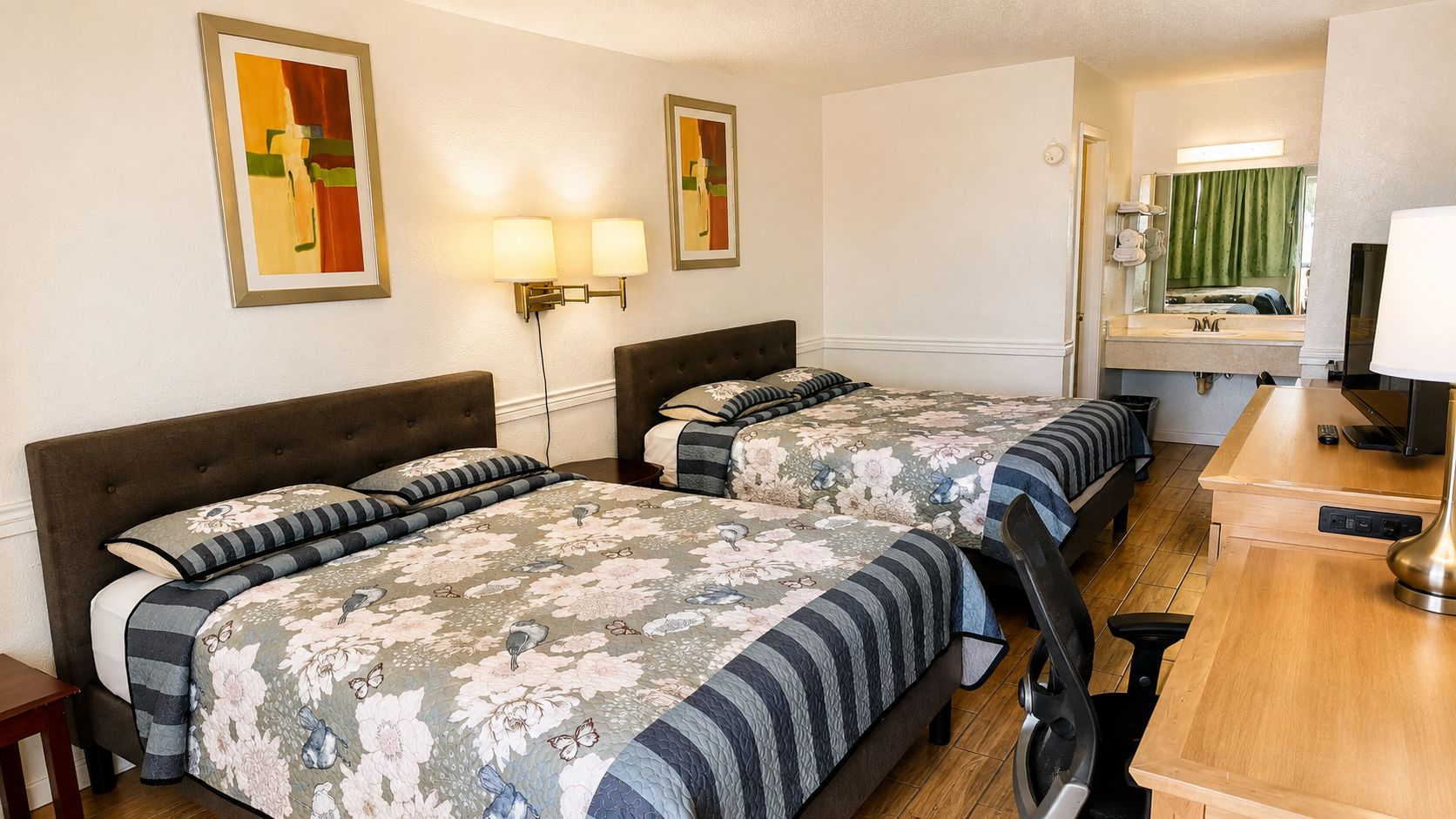 A hotel room with two beds, a desk, a chair, a mirror, and wall-mounted lamps.