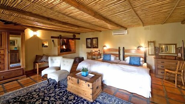 A rustic bedroom with a wooden ceiling, two beds, a wooden chest, and a patterned rug. - De Opstal (Oudtshoorn)