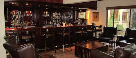 A bar area with multiple stools, a coffee machine, and a variety of bottles on shelves. There are leather chairs and a sofa in the seating area.