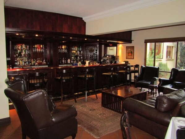Een bar met meerdere krukken, een koffieapparaat en diverse flessen op planken. In het zithoekgedeelte staan leren fauteuils en een bank.
