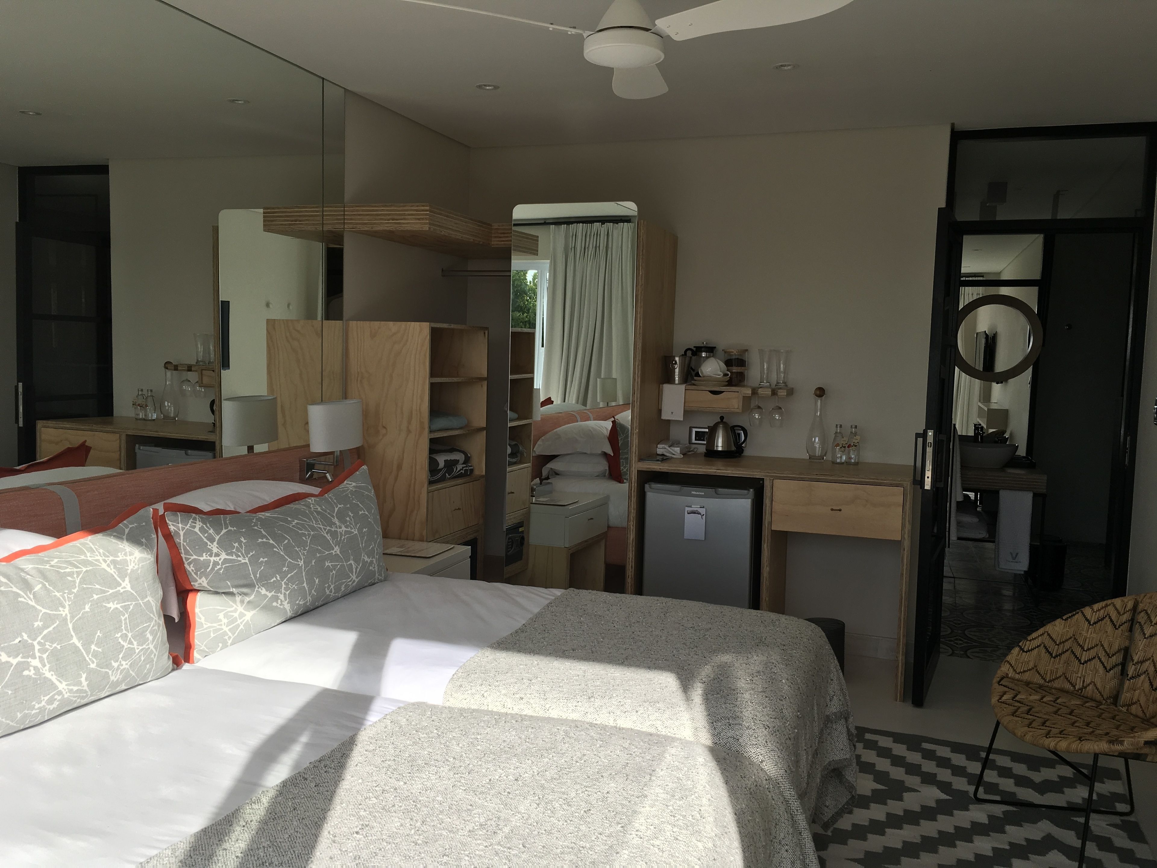 A hotel room with a bed, a desk, a wardrobe, a small fridge, a mirror, and a chair.