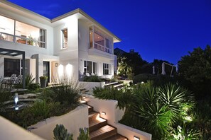 Una casa moderna de dos pisos con un balcón, un jardín bien iluminado y una escalera que conduce al nivel superior.