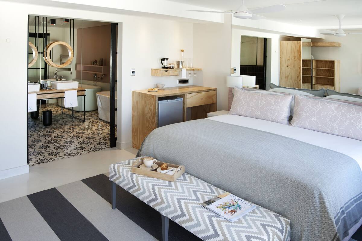 Una habitación de hotel moderna con una cama grande, un escritorio con computadora, un refrigerador pequeño y un baño con espejo y lavamanos.