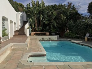 Una piscina con cubierta de baldosas, un conjunto de escaleras que suben a un edificio blanco, y abundante vegetación que rodea el área.