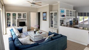 Una sala de estar con un sofá azul, una mesa de centro y un televisor. Hay una zona de cocina con armarios blancos y una ventana con vistas al jardín.
