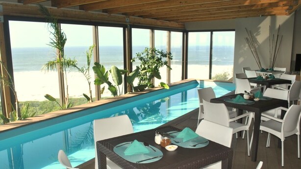 A dining area with a view of the beach and ocean, featuring a pool, white chairs, and a table set with green napkins and condiments. - Wilderness Beach Resort (Wilderness)