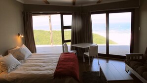 A bedroom with a bed, a desk, and a chair. There are large windows with a view of the outdoors. - Wilderness Beach Resort (Wilderness)