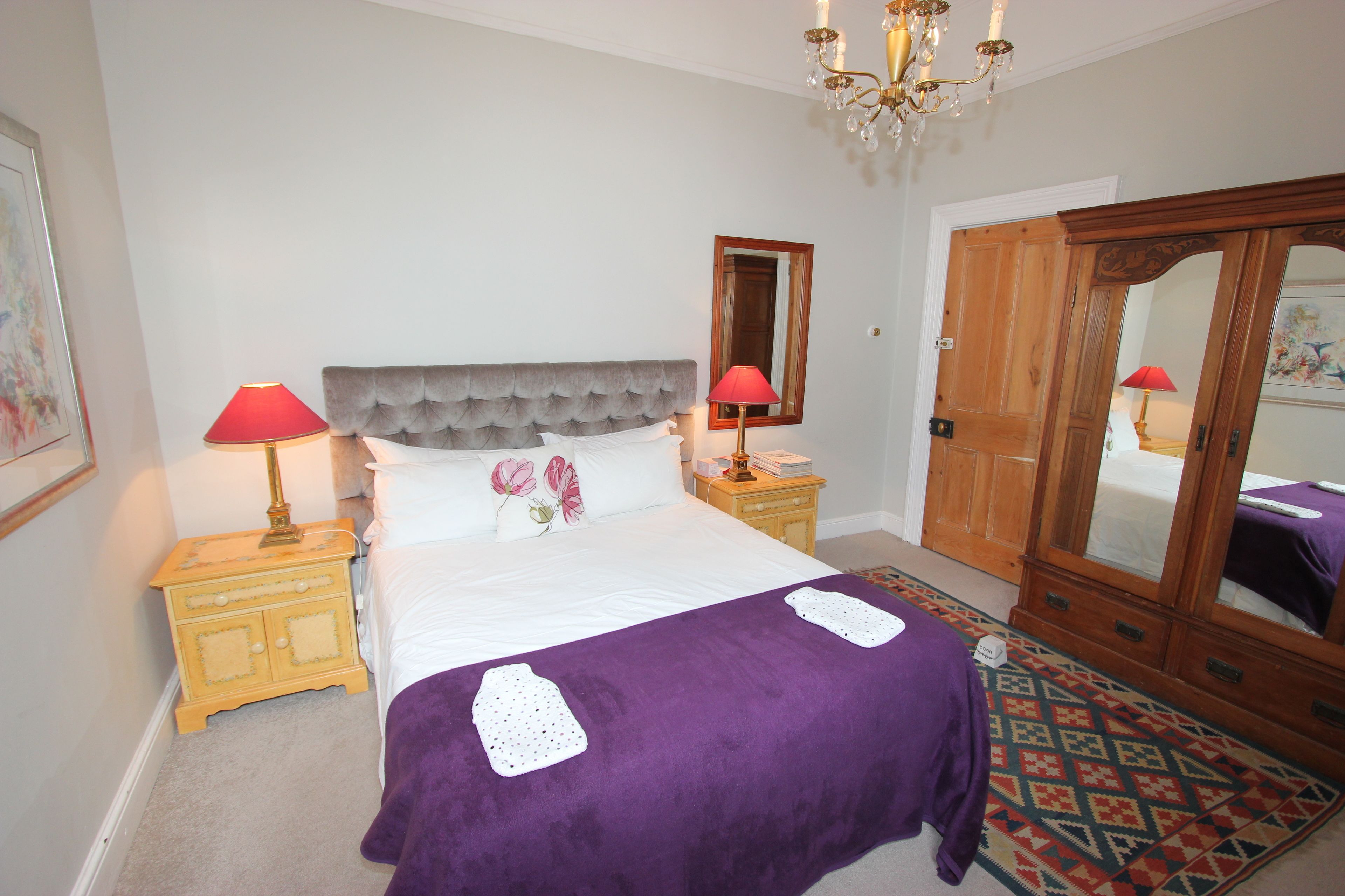A bedroom with a bed, two bedside tables, a nightstand, a chandelier, a mirror, and a wardrobe.