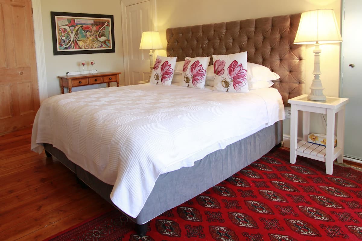 Un dormitorio con una cama grande, un cabecero capitonado, almohadas estampadas con motivos florales, una mesita de noche de madera, una mesita con lámpara y una alfombra roja con diseño.