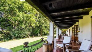 Un espacio al aire libre con techo, con sillas y mesas de madera, rodeado de abundante vegetación y un césped bien cuidado.