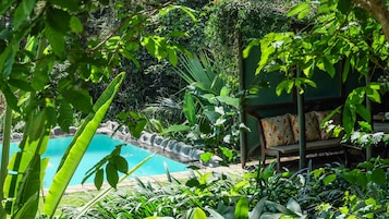 A pool surrounded by lush greenery, a wooden deck, and a seating area with patterned cushions.