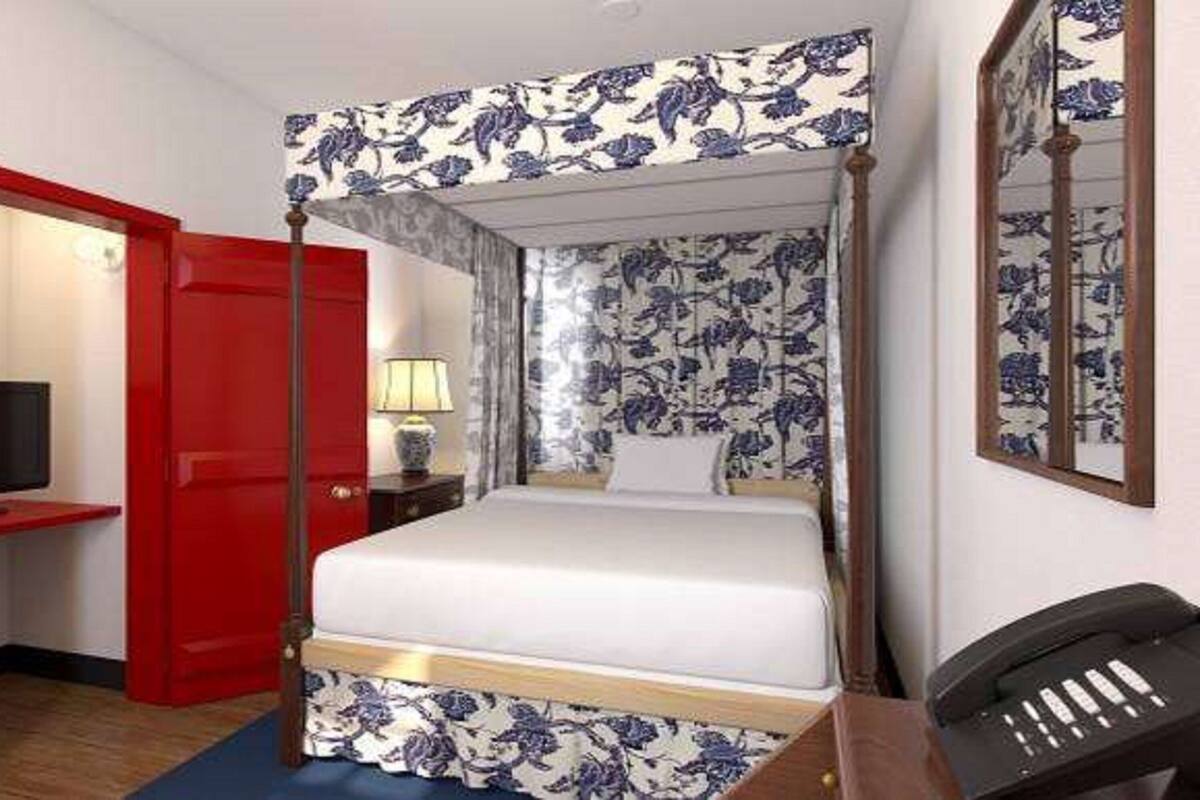 A four-poster bed with floral upholstery, a red door, a bedside table with a lamp, and a telephone.
