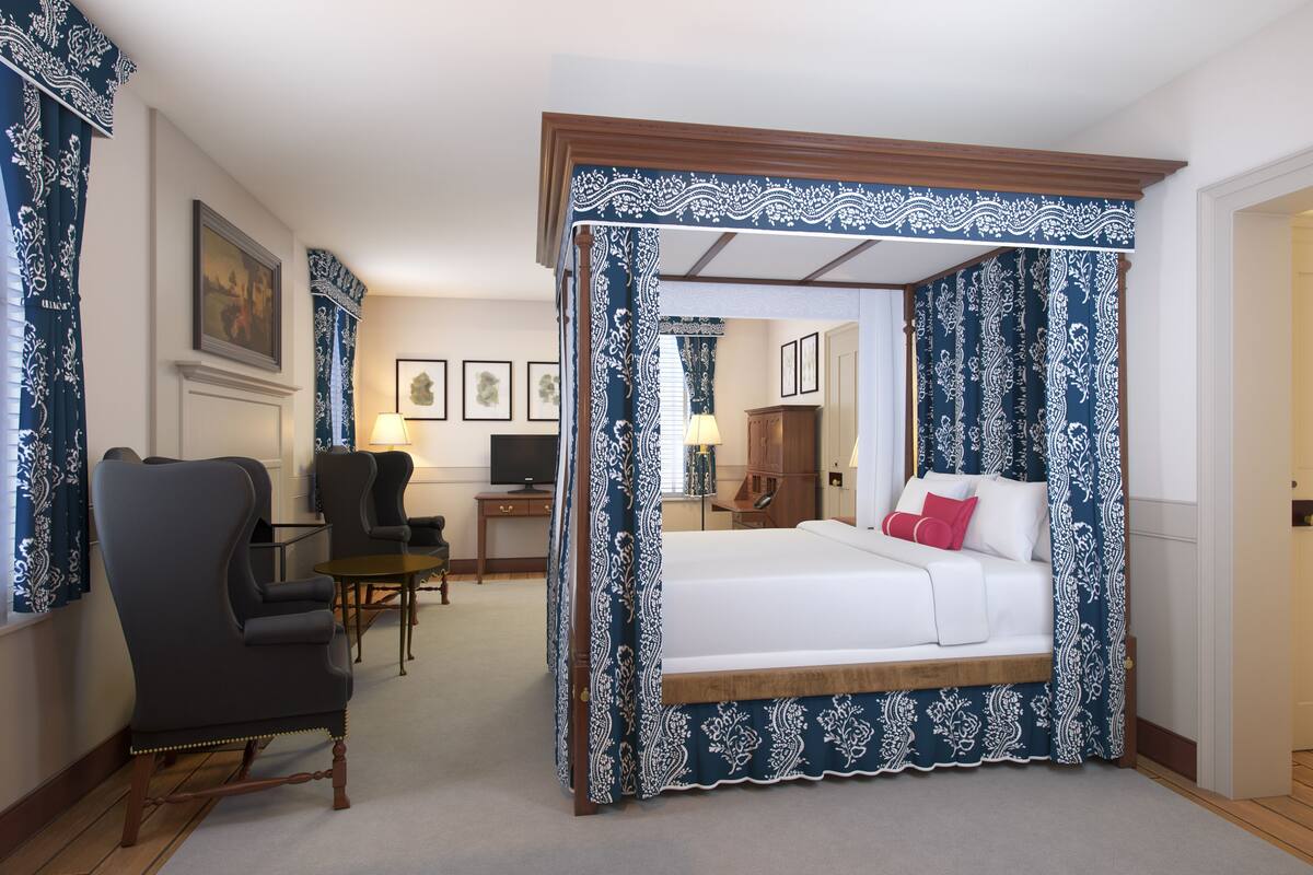 A four-poster canopy bed with a wooden frame and blue and white patterned curtains, a desk with a lamp, and a chair in a hotel room.
