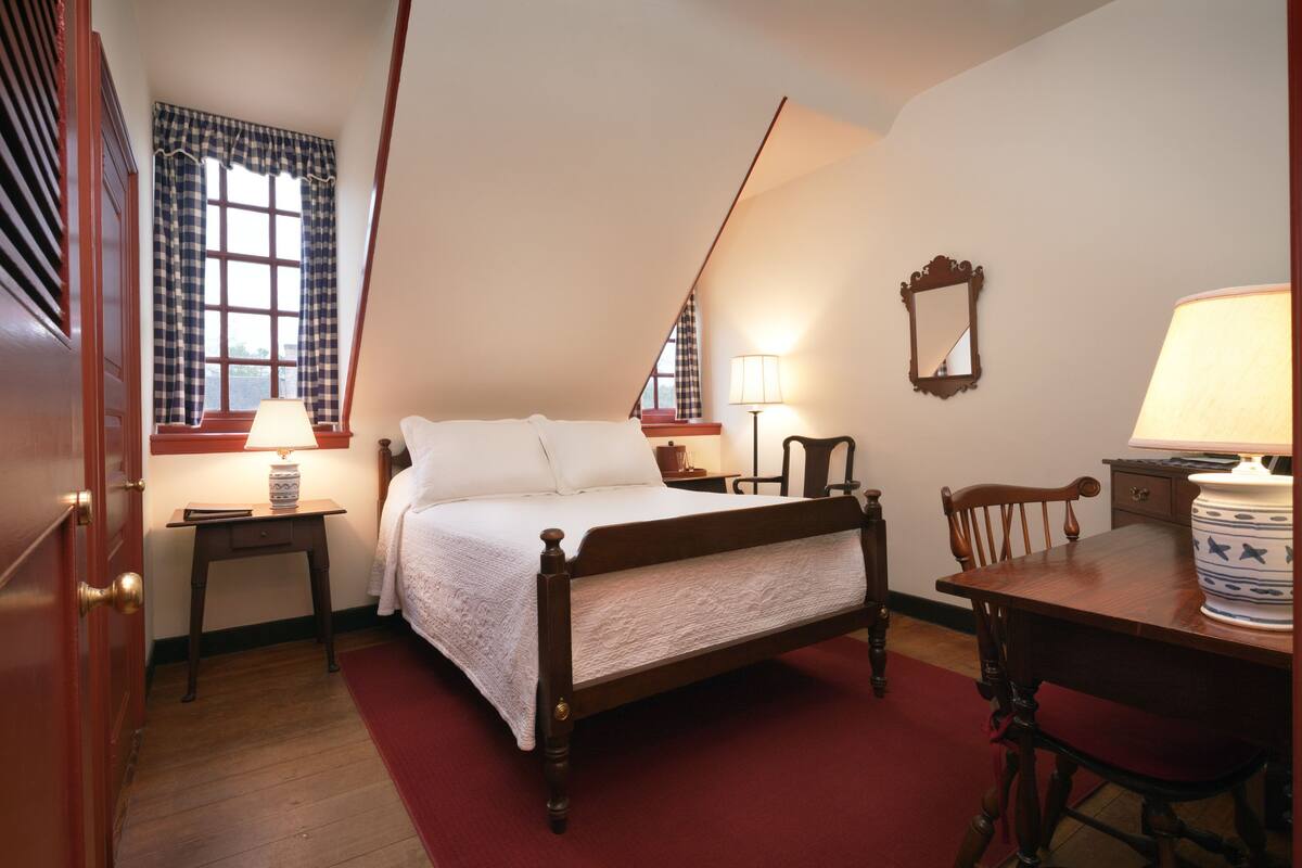 A bedroom with a wooden bed, a desk, a chair, a lamp, a mirror, and a window with curtains.
