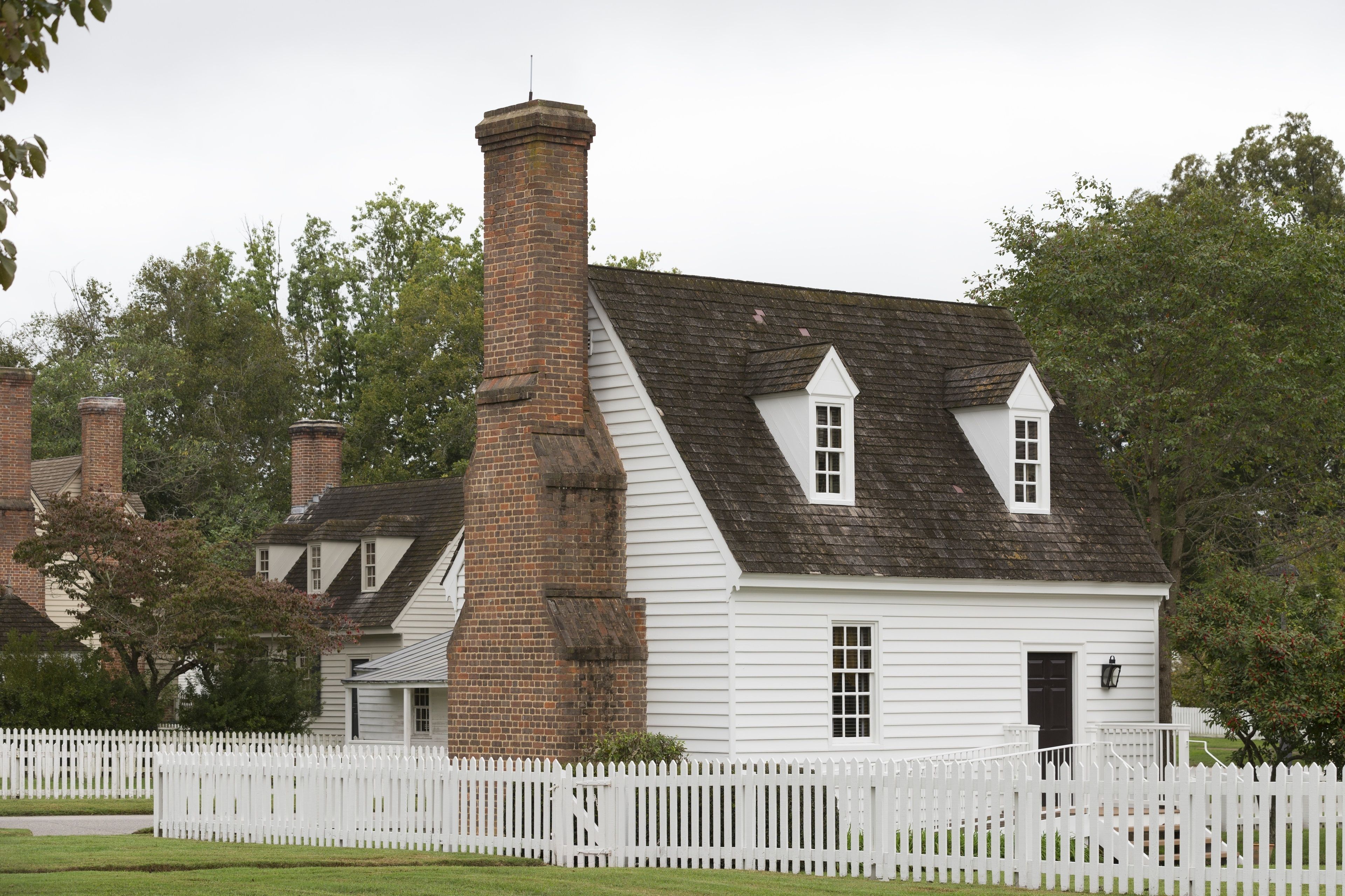 A white colonial-style house with a brick chimney, white picket fence, and a small porch.
