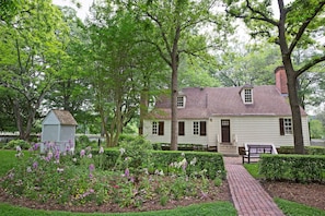 A white colonial-style house with a brick pathway leading to the front door, surrounded by lush greenery and flowering plants. - Colonial Houses, an Official Colonial Williamsburg Hotel (Williamsburg)
