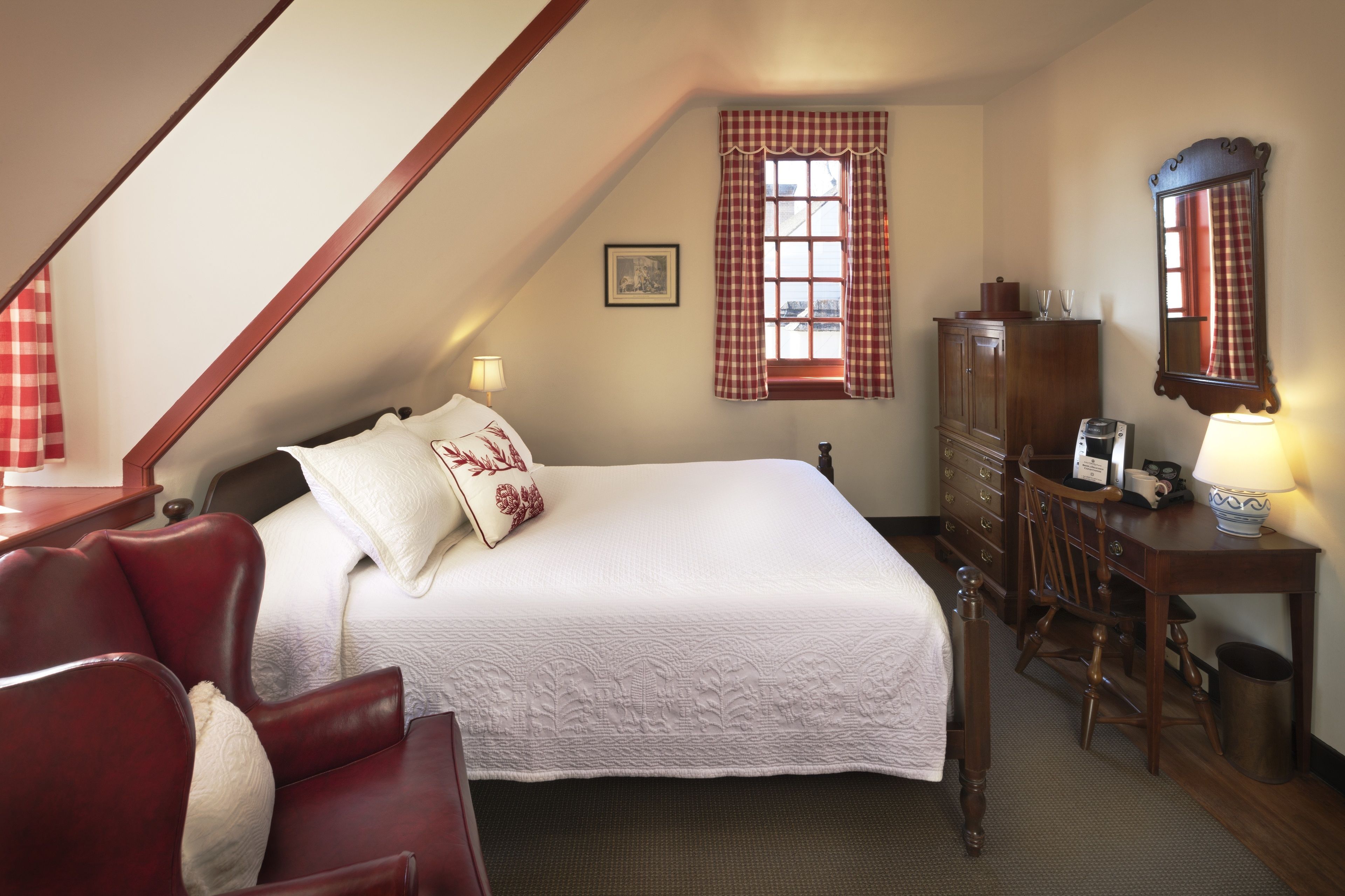 A bedroom with a large bed, a wooden dresser, a desk with a lamp, a chair, and a window with checkered curtains.