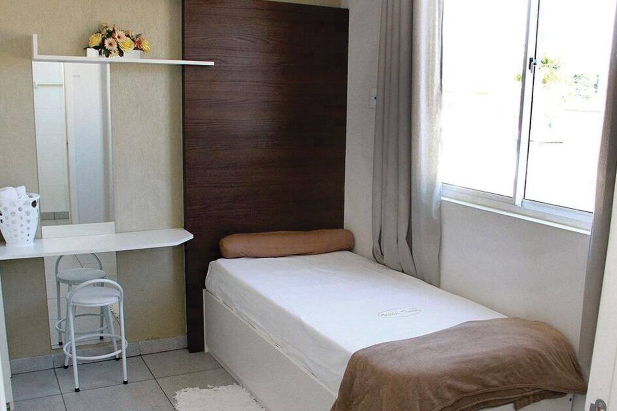 A small, single-bed room with a wooden headboard, a white desk with a chair, a small shelf with flowers, and a window with curtains.