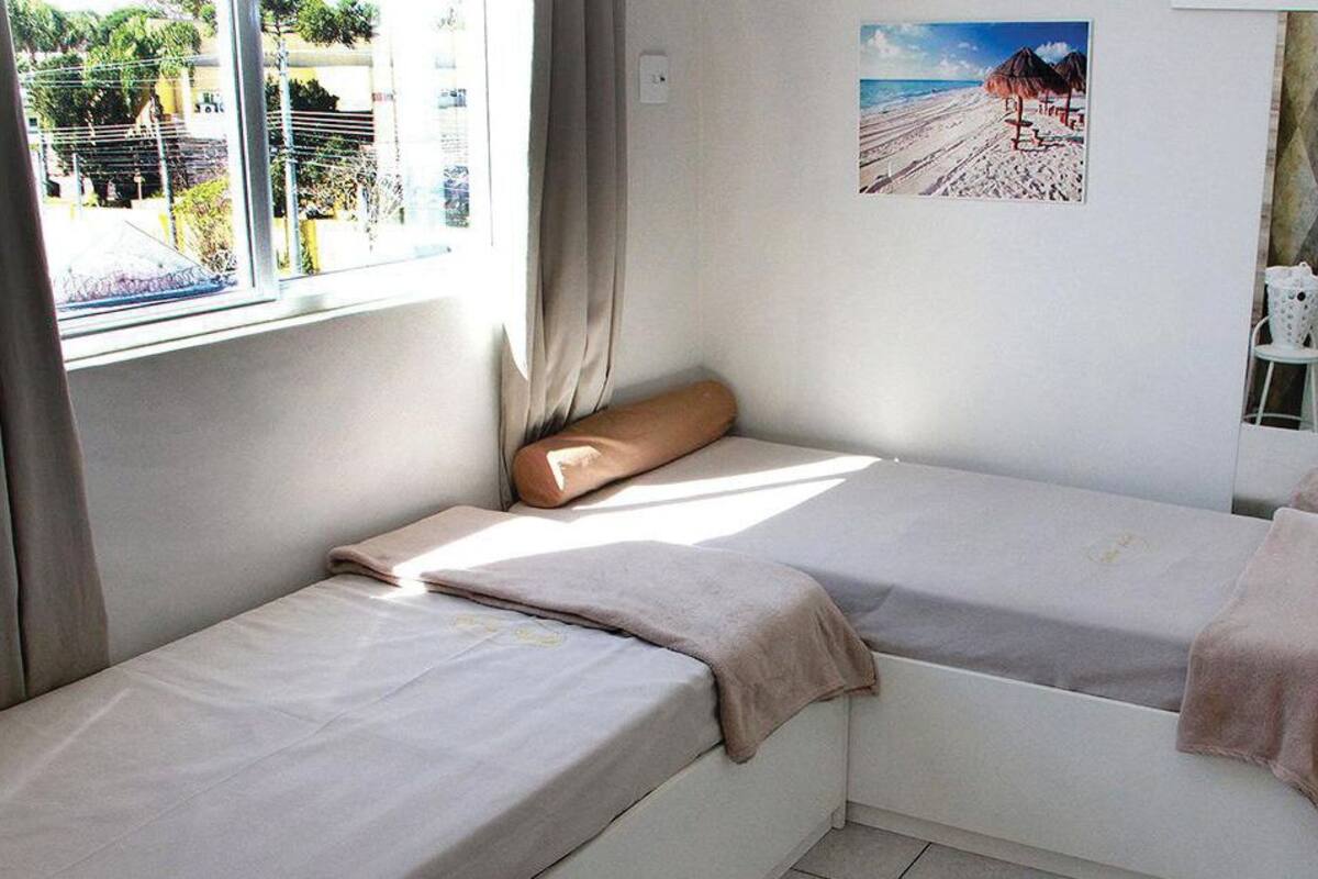 A room with two beds, a window with curtains, a beach picture on the wall, and a chair with a bag.