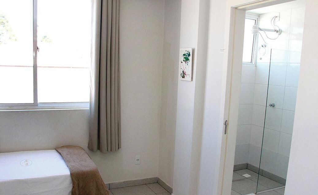 A small, clean room with a bed, a window with curtains, a shower area with glass doors, and a wall-mounted artwork.