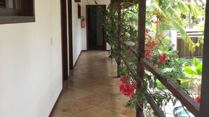 A covered corridor with tiled flooring, wooden beams, and a view of a garden with red flowers and a parked car. - Pousada Point Break (Caucaia)